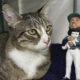 A tabby cat sits beside a baseball player bobblehead holding a small white cat plush, resting on a blue blanket in a red tub corner.