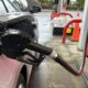 Close-up of a gas pump nozzle inserted into a silver car's fuel filler at a fueling station, with orange cones and a red Fred Meyer sign in the background.