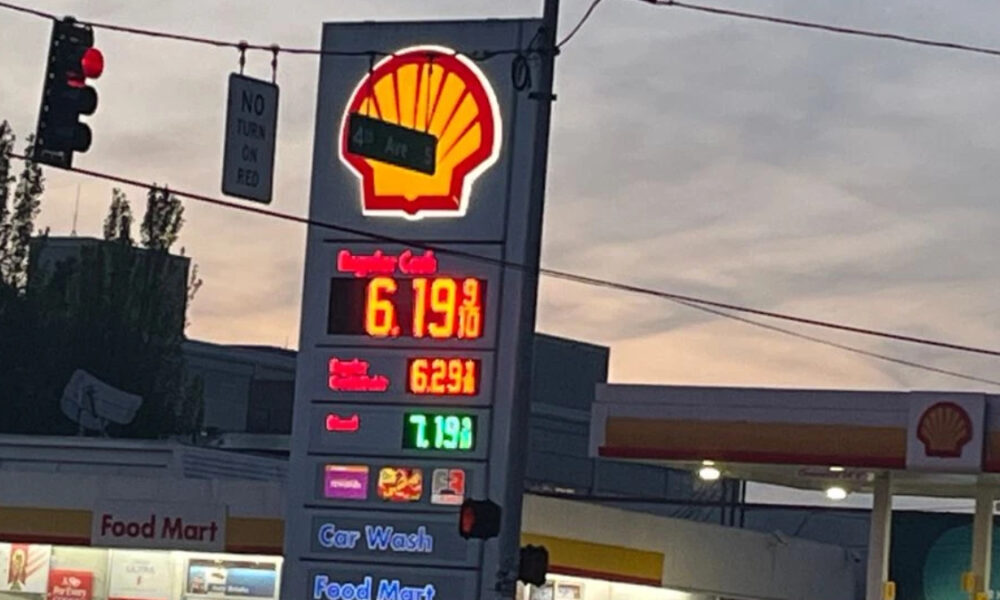 Shell gas station with a tall illuminated price board showing fuel prices; red/yellow logo at top, nearby Food Mart storefront and a car wash sign visible at dusk.