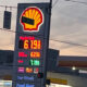 Shell gas station with a tall illuminated price board showing fuel prices; red/yellow logo at top, nearby Food Mart storefront and a car wash sign visible at dusk.