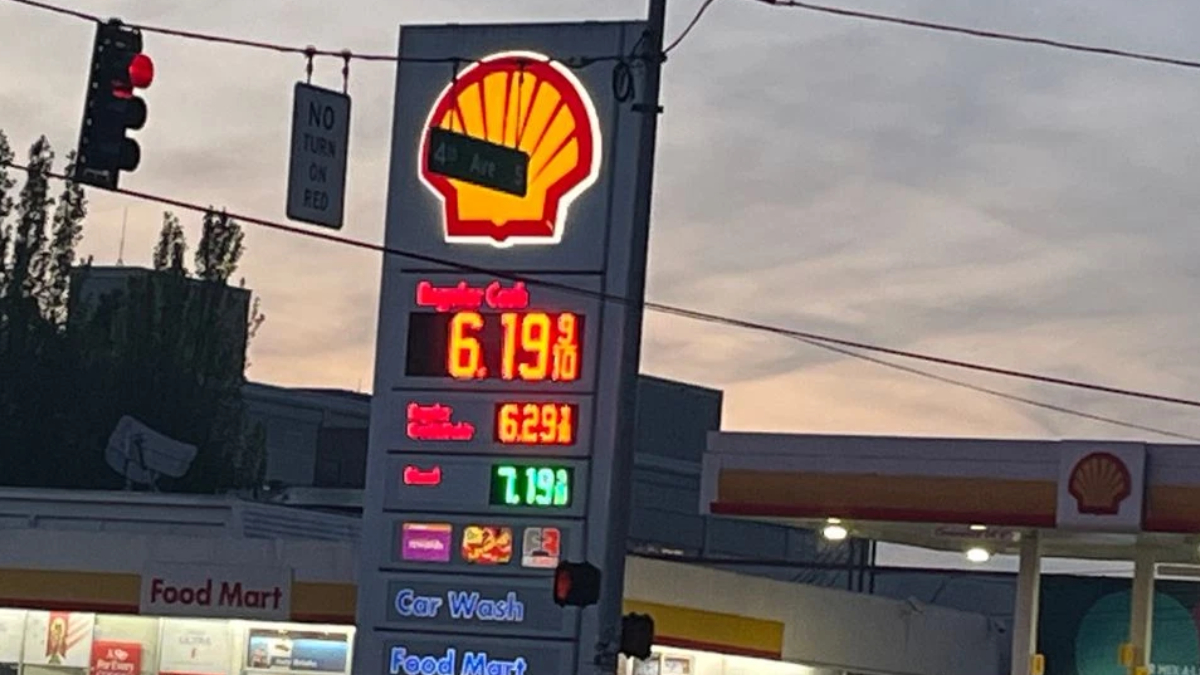 Shell gas station with a tall illuminated price board showing fuel prices; red/yellow logo at top, nearby Food Mart storefront and a car wash sign visible at dusk.