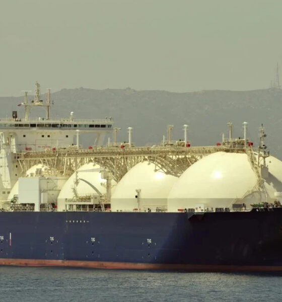 Large blue LNG carrier with three white spherical tanks on calm sea, hills in the background.