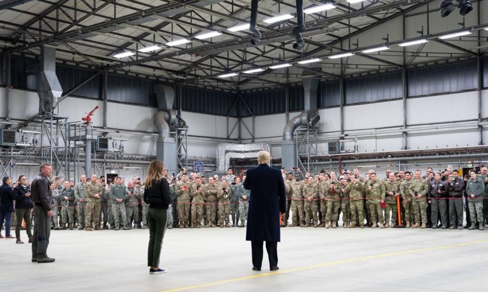 A speaker in a dark coat addresses a large group of soldiers lined up inside a spacious industrial hangar.