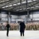A speaker in a dark coat addresses a large group of soldiers lined up inside a spacious industrial hangar.