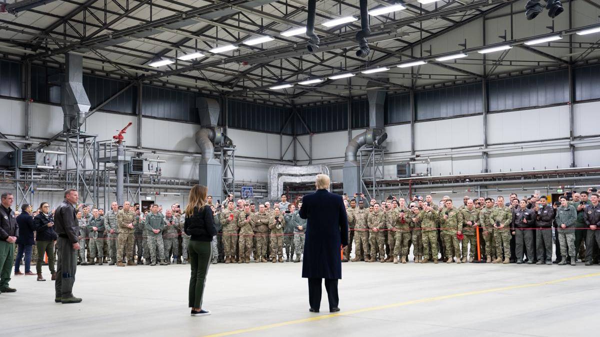 A speaker in a dark coat addresses a large group of soldiers lined up inside a spacious industrial hangar.