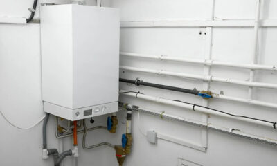 White wall-mounted boiler with exposed pipes, valves, and conduit in a utility room.