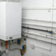 White wall-mounted boiler with exposed pipes, valves, and conduit in a utility room.
