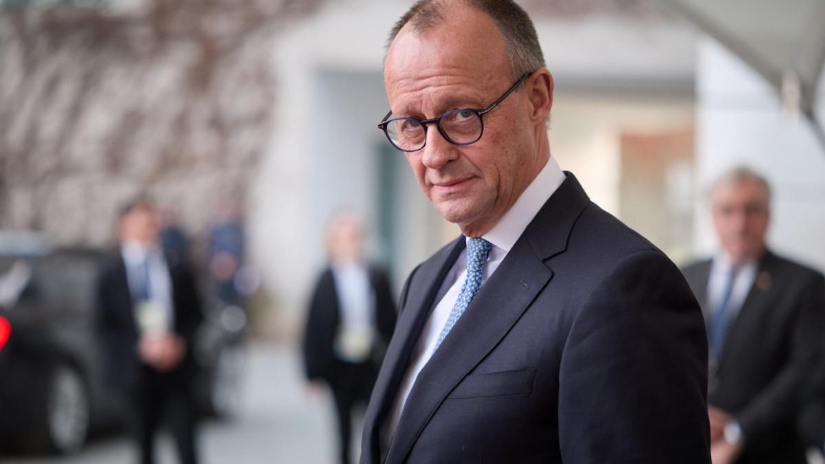 A bespectacled businessman in a dark suit and blue tie looking toward the camera outdoors, with blurred people in the background.