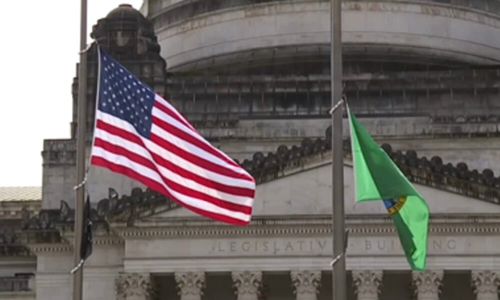 American flag and a green flag waving in front of a neoclassical legislative building