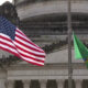 American flag and a green flag waving in front of a neoclassical legislative building
