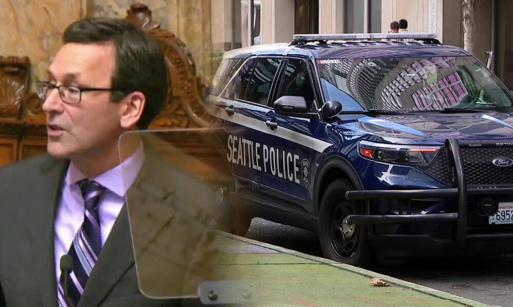 Man in a suit speaks at a podium with a transparent shield as a Seattle Police SUV sits parked on a city street in the background.