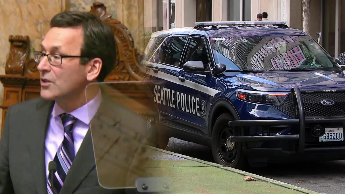 Man in a suit speaks at a podium with a transparent shield as a Seattle Police SUV sits parked on a city street in the background.