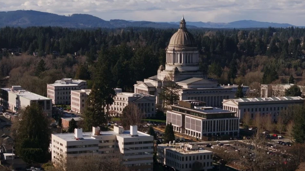 Aerial view of a domed government building surrounded by modern office blocks and trees in a cityscape.