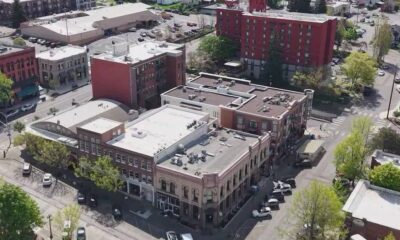 Aerial view of a small downtown block with brick buildings, trees, and parked cars along intersecting streets. Red brick building stands out in the center-right.