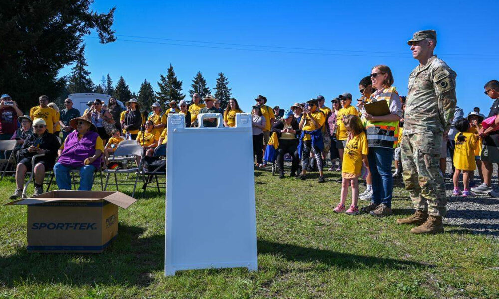 Crowd of people at an outdoor community event, many in yellow shirts, with a soldier and a organizer nearby under a clear blue sky.