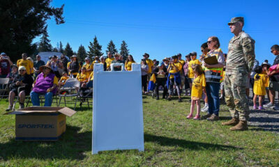 Crowd of people at an outdoor community event, many in yellow shirts, with a soldier and a organizer nearby under a clear blue sky.