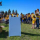 Crowd of people at an outdoor community event, many in yellow shirts, with a soldier and a organizer nearby under a clear blue sky.