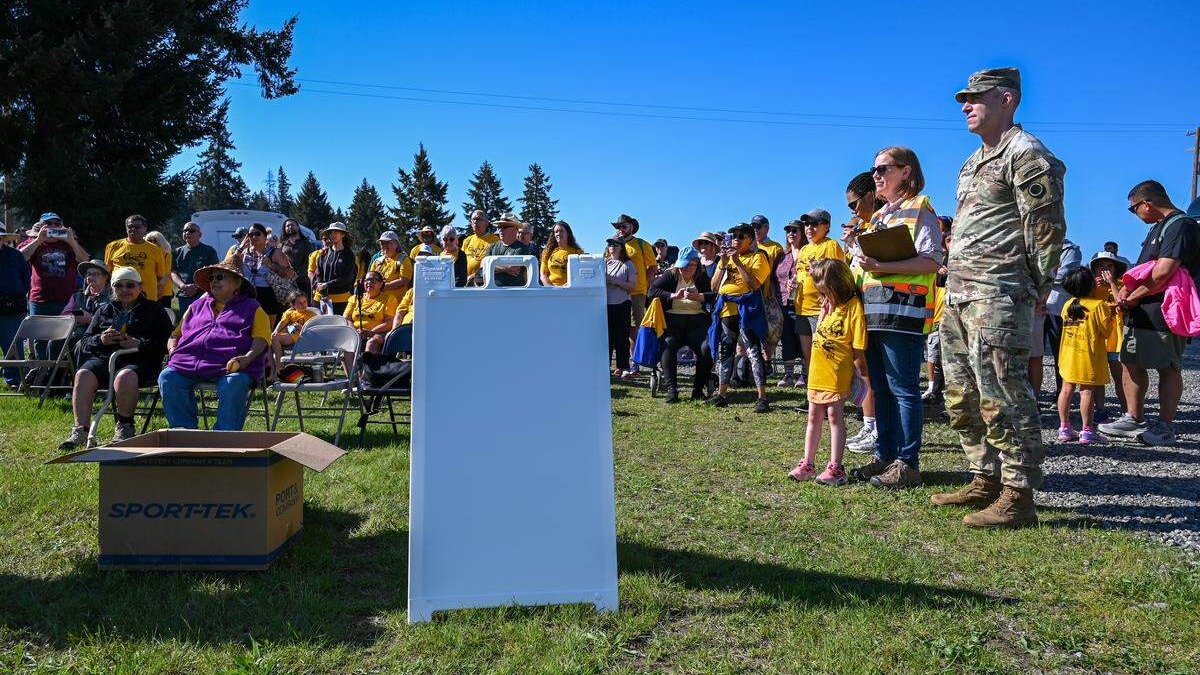 Crowd of people at an outdoor community event, many in yellow shirts, with a soldier and a organizer nearby under a clear blue sky.