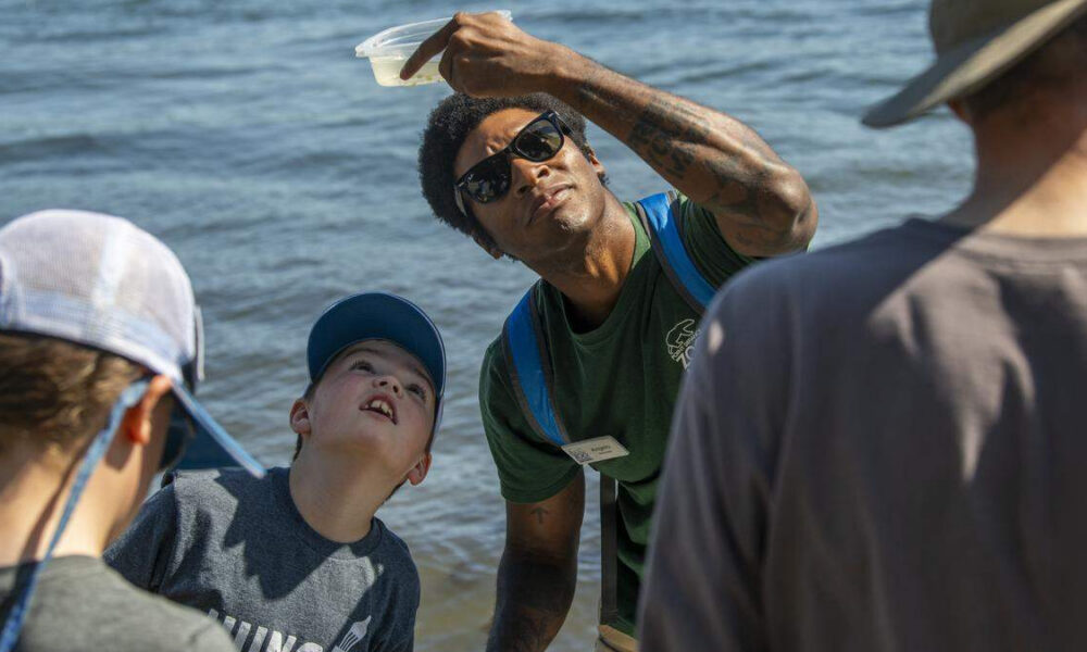 Instructor with sunglasses holds up a clear plastic bag of water for kids by the lake shore.