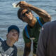 Instructor with sunglasses holds up a clear plastic bag of water for kids by the lake shore.