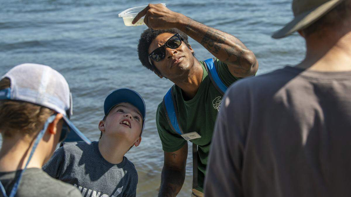 Instructor with sunglasses holds up a clear plastic bag of water for kids by the lake shore.