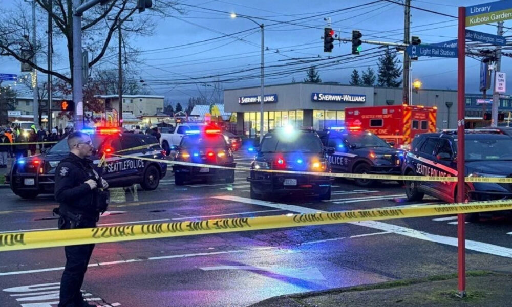 Police cars with flashing red and blue lights block an intersection, yellow caution tape across the street, bystanders in the background, an ambulance nearby, and a Sherwin-Williams store in view.