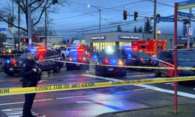 Police cars with flashing red and blue lights block an intersection, yellow caution tape across the street, bystanders in the background, an ambulance nearby, and a Sherwin-Williams store in view.