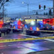 Police cars with flashing red and blue lights block an intersection, yellow caution tape across the street, bystanders in the background, an ambulance nearby, and a Sherwin-Williams store in view.