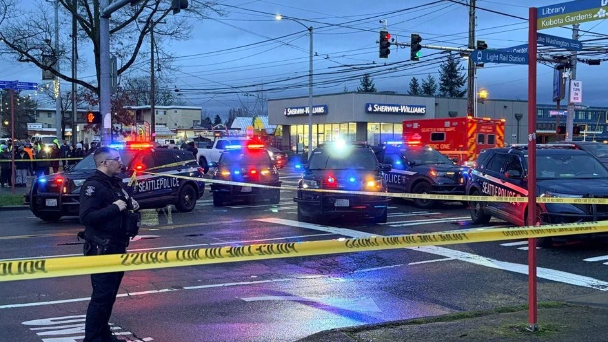 Police cars with flashing red and blue lights block an intersection, yellow caution tape across the street, bystanders in the background, an ambulance nearby, and a Sherwin-Williams store in view.