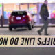 Crime scene tape in the foreground reads 'DO NOT CROSS' (inverted); blurred man and a red SUV seen near a storefront.