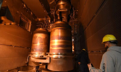 Two workers in yellow hard hats observe large copper-colored industrial vessels mounted in a metal-walled chamber, warm orange lighting surrounding the equipment.
