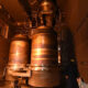 Two workers in yellow hard hats observe large copper-colored industrial vessels mounted in a metal-walled chamber, warm orange lighting surrounding the equipment.