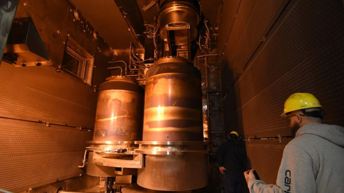 Two workers in yellow hard hats observe large copper-colored industrial vessels mounted in a metal-walled chamber, warm orange lighting surrounding the equipment.