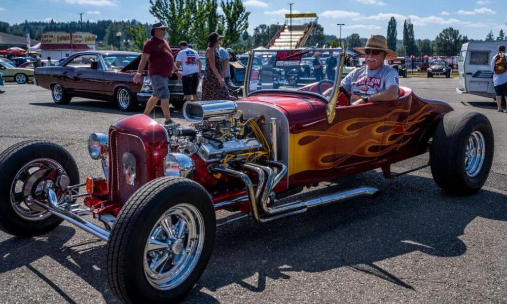 Red flame-painted vintage hot rod with an exposed engine at a car show, with people strolling in the background.
