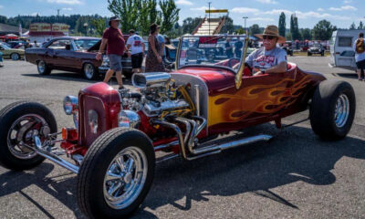 Red flame-painted vintage hot rod with an exposed engine at a car show, with people strolling in the background.