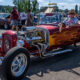 Red flame-painted vintage hot rod with an exposed engine at a car show, with people strolling in the background.