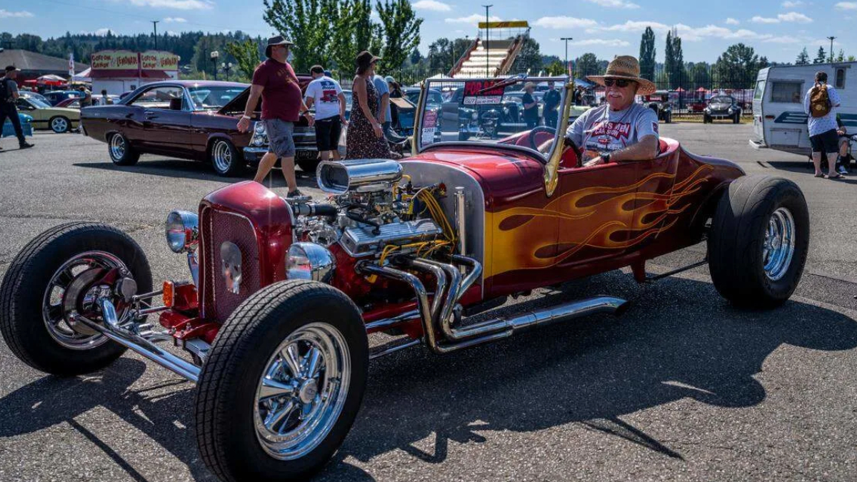 Red flame-painted vintage hot rod with an exposed engine at a car show, with people strolling in the background.