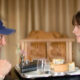 A man wearing a blue cap leans toward a woman across a table, smiling, with a plate of cake and a glass between them on a dining tray.