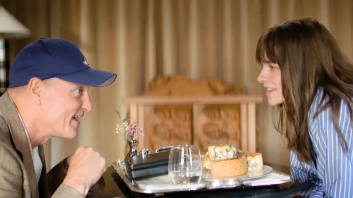 A man wearing a blue cap leans toward a woman across a table, smiling, with a plate of cake and a glass between them on a dining tray.