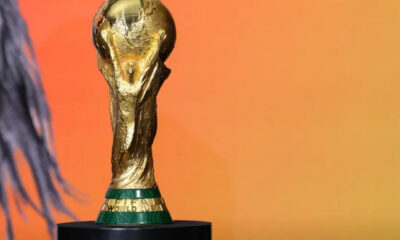 FIFA World Cup trophy in gold on a black pedestal against an orange gradient background.