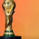 FIFA World Cup trophy in gold on a black pedestal against an orange gradient background.