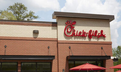 Chick-fil-A restaurant exterior with red logo on a beige brick wall and red umbrellas outside.