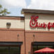 Chick-fil-A restaurant exterior with red logo on a beige brick wall and red umbrellas outside.