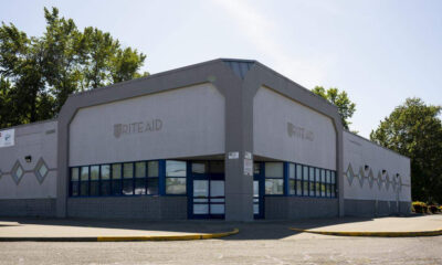 Front view of a Rite Aid store with gray facade, blue-framed windows, and a closed handicap curb; empty parking lot under a clear sky.