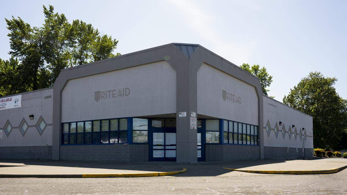 Front view of a Rite Aid store with gray facade, blue-framed windows, and a closed handicap curb; empty parking lot under a clear sky.