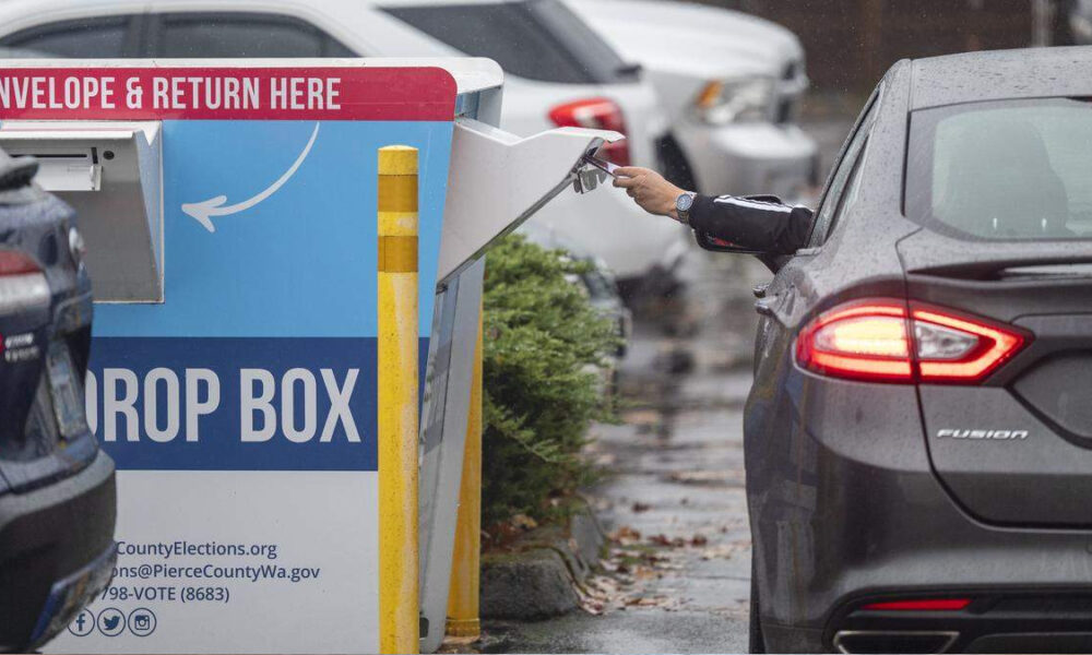 Driver inserts a ballot into a blue drop box labeled DROP BOX in a parking lot, with a car nearby on a rainy day.