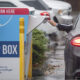 Driver inserts a ballot into a blue drop box labeled DROP BOX in a parking lot, with a car nearby on a rainy day.