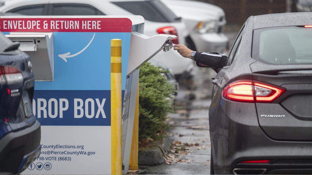 Driver inserts a ballot into a blue drop box labeled DROP BOX in a parking lot, with a car nearby on a rainy day.