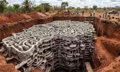 Deep excavation at a construction site with a massive stack of interlocking concrete forms in the trench; workers stand near the edge in the background.