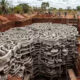 Deep excavation at a construction site with a massive stack of interlocking concrete forms in the trench; workers stand near the edge in the background.